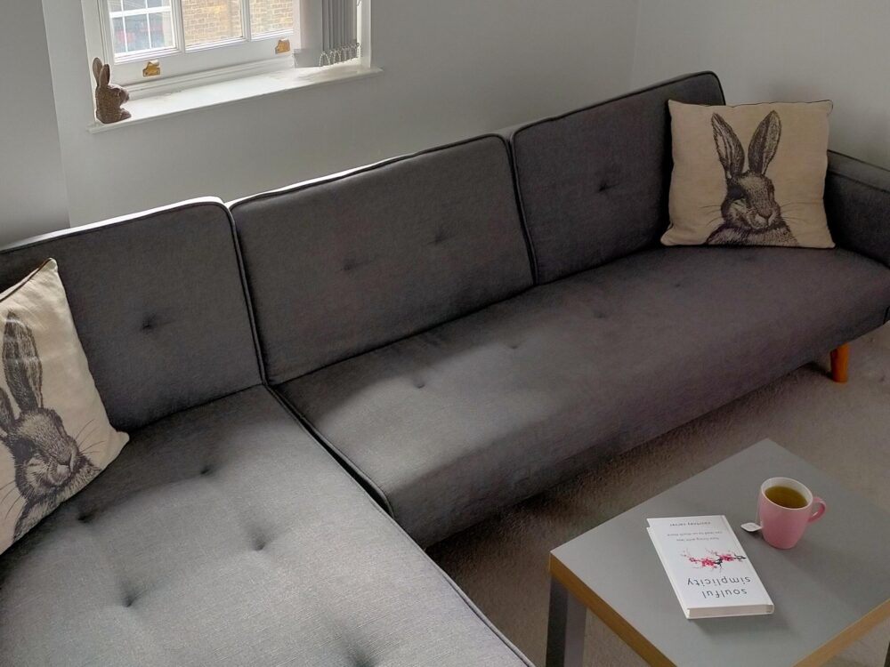 Living room with large grey corner sofa with cushions with rabbit print, and a coffee table with a cup of tea and the book Soulful Simplicity