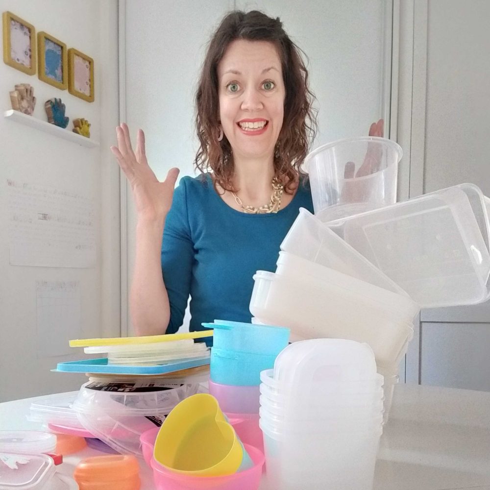 Woman sitting at the table with her hands up in the air in a desperate manner, with a mock desperate look on her face, with lots of plastic (tupperware) containers in front of her.